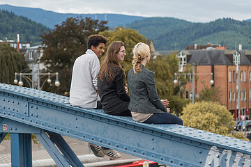 Drei Studierenden unterschiedlicher Herkünfte sitzen auf der Blauen Brücke in Freiburg